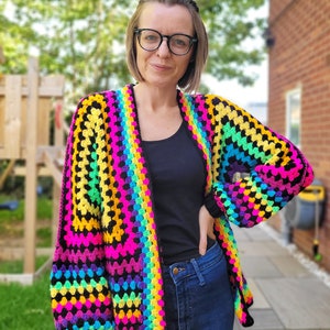 K&ouml;nnte beinhalten: Eine Regenbogen-H&auml;keljacke mit schwarzem Hintergrund und langen &Auml;rmeln. Die Jacke ist vorne offen und hat eine lockere Passform.