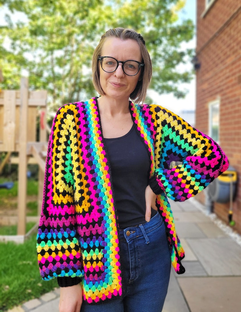 K&ouml;nnte beinhalten: Eine Regenbogen-H&auml;keljacke mit schwarzem Hintergrund und langen &Auml;rmeln. Die Jacke ist vorne offen und hat eine lockere Passform.