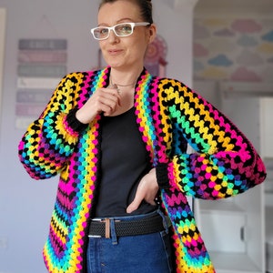 May include: A rainbow crochet cardigan with black stripes. The cardigan is open in the front and has long sleeves. The person wearing the cardigan is also wearing a black shirt and blue jeans.