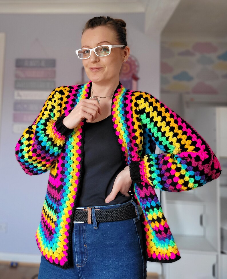 May include: A rainbow crochet cardigan with black stripes. The cardigan is open in the front and has long sleeves. The person wearing the cardigan is also wearing a black shirt and blue jeans.