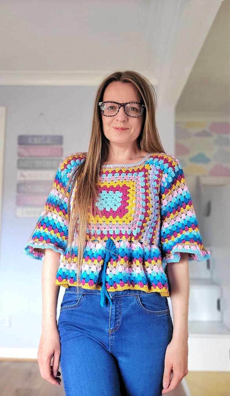 May include: A colorful crocheted top with a square design in the center and a tie at the waist. The top is worn with blue jeans.