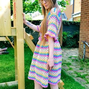 May include: A woman wearing a colorful crochet dress with a striped pattern. The dress has a bell sleeve and a crochet trim. The woman is standing in a garden.