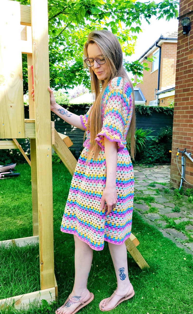 May include: A woman wearing a colorful crochet dress with a striped pattern. The dress has a bell sleeve and a crochet trim. The woman is standing in a garden.