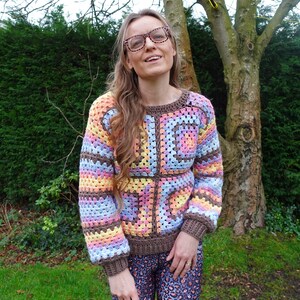 May include: A woman wearing a colorful crocheted sweater with a brown background and a geometric pattern. The sweater has long sleeves and a relaxed fit.