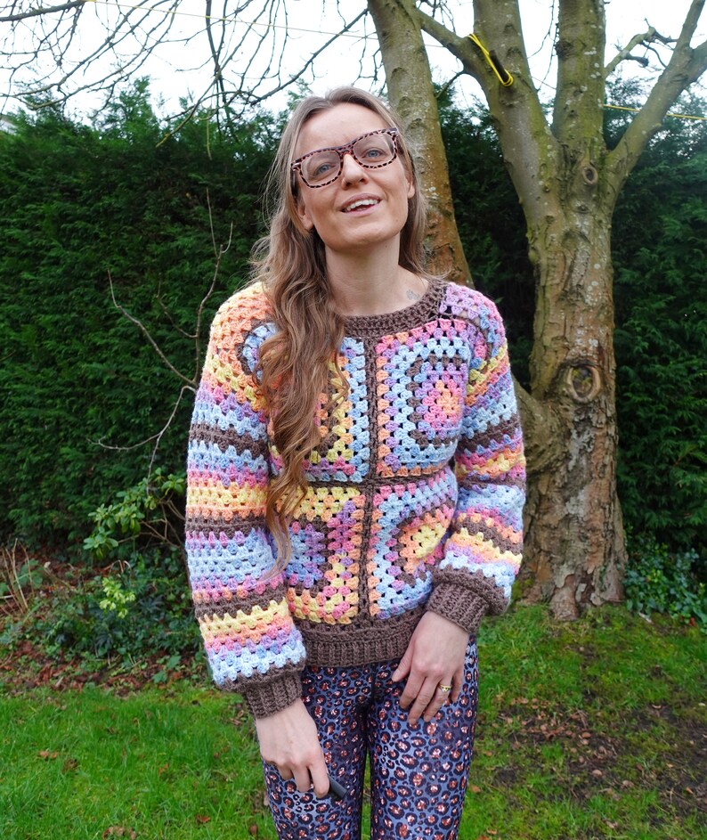 May include: A woman wearing a colorful crocheted sweater with a brown background and a geometric pattern. The sweater has long sleeves and a relaxed fit.