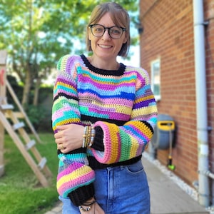 May include: A woman wearing a colorful striped crochet sweater with long sleeves and a round neckline. She is wearing blue denim shorts and has glasses on.
