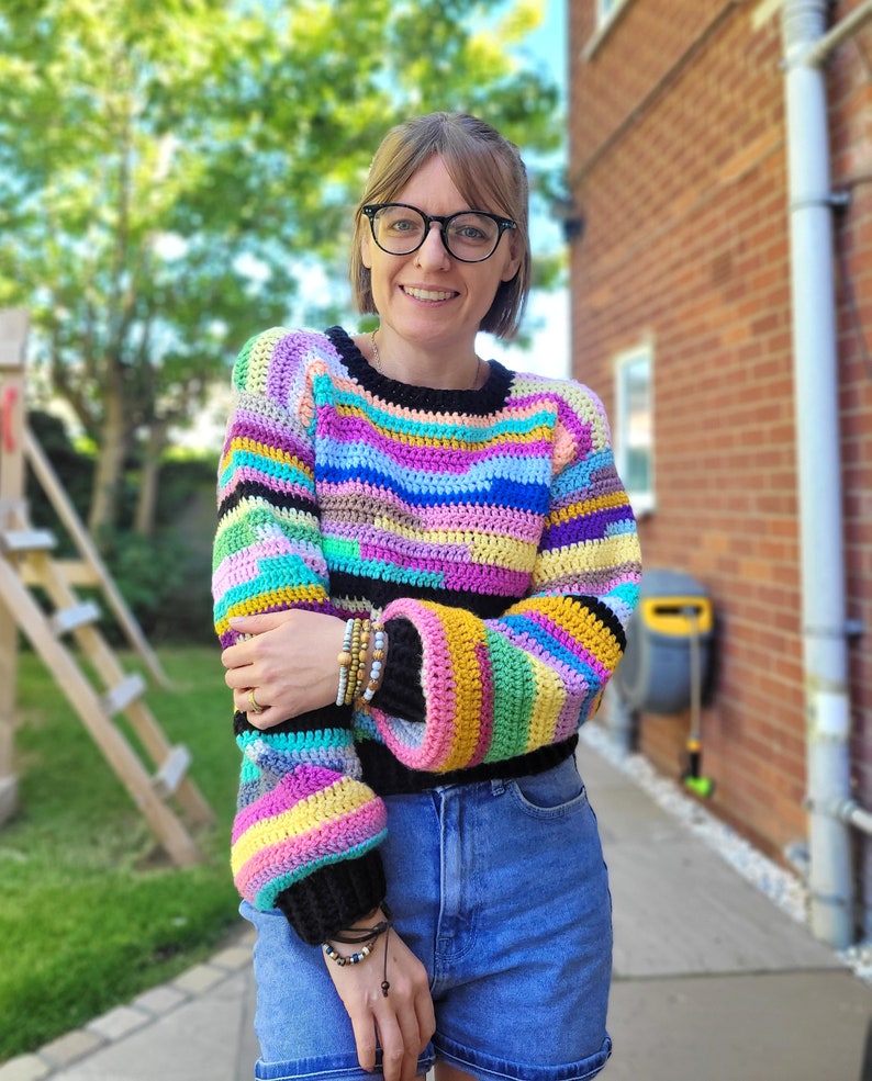 May include: A woman wearing a colorful striped crochet sweater with long sleeves and a round neckline. She is wearing blue denim shorts and has glasses on.