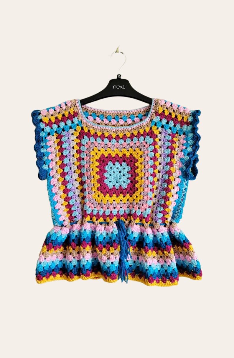 May include: A colorful crochet top with a square pattern and a ruffled bottom. The top is made with a variety of colors, including pink, blue, yellow, and green. It has a tassel tie at the front.