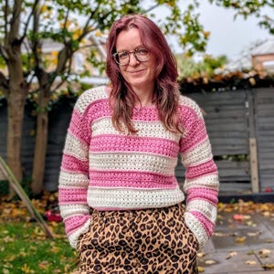 Könnte beinhalten: Ein rosa-weiß gestreifter Häkelpullover mit langen Ärmeln. Der Pullover wird mit einer Hose mit Leopardenmuster kombiniert. Die Person trägt eine Brille und steht draußen vor einem Zaun und Bäumen.