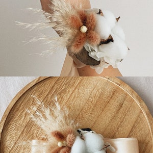 May include: A wrist corsage made with dried pampas grass, cotton bolls, and a dried flower. The corsage is tied with a white satin ribbon and is displayed on a wooden plate.