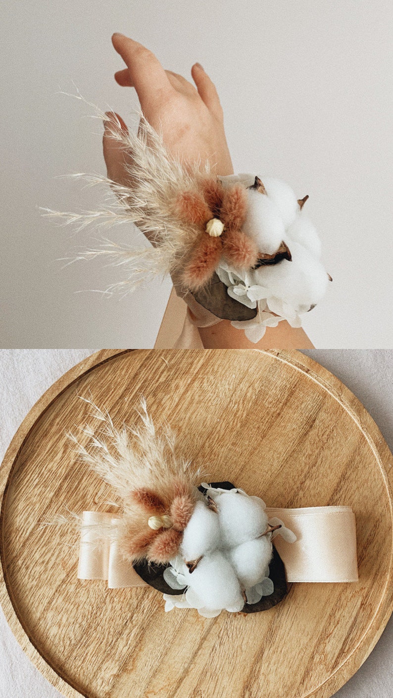 May include: A wrist corsage made with dried pampas grass, cotton bolls, and a dried flower. The corsage is tied with a white satin ribbon and is displayed on a wooden plate.