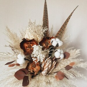 May include: A dried flower bouquet with white pampas grass, brown feathers, dried eucalyptus leaves, and cotton bolls.