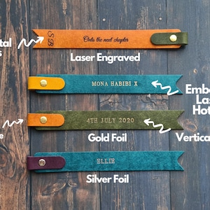 May include: Four leather bookmarks with different personalization options. The top bookmark is laser engraved with the initials "SB" and the text "Onto the next chapter". The second bookmark is embossed with the text "MONA HABIBI X". The third bookmark is embossed with the text "4TH JULY 2020" in gold foil. The bottom bookmark is embossed with the name "ELLIE" in silver foil.