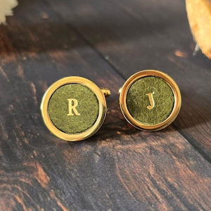 May include: Two gold-toned cufflinks with olive green centers. One cufflink displays the letter "R", the other the letter "J". The cufflinks are set against a dark wood grain background, with decorative elements in the periphery.