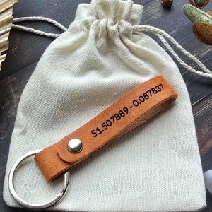 May include: A tan leather keyring featuring a silver metal ring and a silver rivet, with the coordinates 51.507889 -0.087837 embossed in black on the leather strap, resting on a small, off-white drawstring canvas pouch.