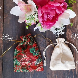 May include: Three drawstring bags on a wooden surface. The bags are made of different materials: a red and green Christmas-themed bag, a plain white cotton bag, and a natural-colored cotton bag.