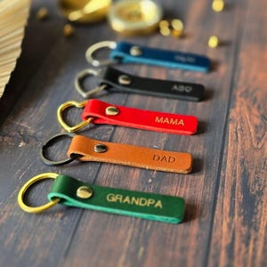 Könnte beinhalten: Satz von sechs Leder-Schlüsselanhängern mit verschiedenen farbigen Lederriemen und Metallringen. Die Riemen sind mit den Worten "Grandpa", "Dad", "Mama", "Abo" und zwei weiteren Wörtern graviert, die auf dem Bild nicht klar zu erkennen sind.