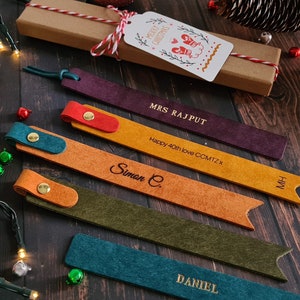 May include: Five colorful leather bookmarks with personalized names, including "Mrs. Rajput", "Simon C.", "Daniel", "MH", and "Happy 40th Love CCMTZX". The bookmarks are arranged on a wooden surface with Christmas decorations.