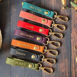 May include: Eight leather keyrings with gold metal key rings. Each keyring has a different colour and is personalised with a name or phrase, including "Love", "Grandma", "Grandpa", "Daddy", "Flat 2", "Mum", "Na Oer".
