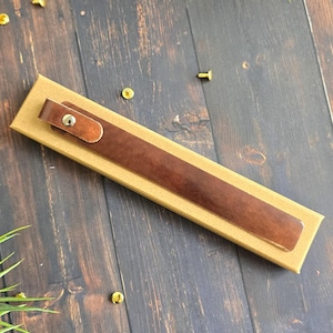May include: A brown leather pen sleeve with a snap closure, resting on a rectangular cardboard box. The pen sleeve is a rich, dark brown color. The box is a light tan color. The background is a dark wood surface.
