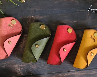 Könnte beinhalten: Vier Leder-Schlüsselanhänger in verschiedenen Farben, jeder mit einem personalisierten Monogramm. Die Schlüsselanhänger sind in der Mitte gefaltet und haben einen goldenen Knopfverschluss. Die Farben sind rosa, grün, rot und gelb.