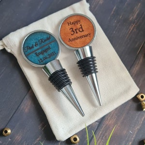 May include: Two personalised bottle stoppers with engraved text. One stopper has a teal blue background with the text "Paul & Rachel Engaged 12.06.2021". The other stopper has an orange background with the text "Happy 3rd Anniversary".
