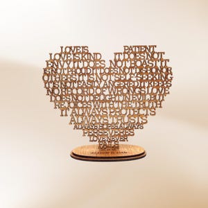 May include: A wooden heart-shaped decorative piece with the words "Love is Kind" and other phrases laser-cut into the design. The piece sits on a round wooden base with the names "Allison & Dan" engraved.