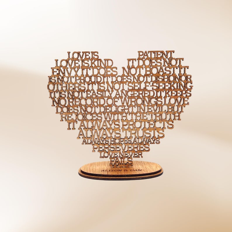 May include: A wooden heart-shaped decorative piece with the words "Love is Kind" and other phrases laser-cut into the design. The piece sits on a round wooden base with the names "Allison & Dan" engraved.