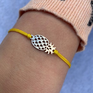 May include: A yellow cord bracelet featuring a silver pineapple charm. The charm has a cut-out design. The bracelet is worn on a wrist, against a peach-coloured knitted sleeve.