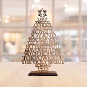 以下が含まれることがあります： 木製のクリスマスツリーの装飾品。テキスト「MAY THE GOD OF HOPE FILL YOU WITH ALL JOY AND PEACE AS YOU TRUST IN HIM SO THAT YOU MAY OVERFLOW WITH HOPE BY THE POWER OF THE HOLY SPIRIT ROMANS 15:13」が刻まれています。茶色の木製で、濃い茶色の台座に立っています。