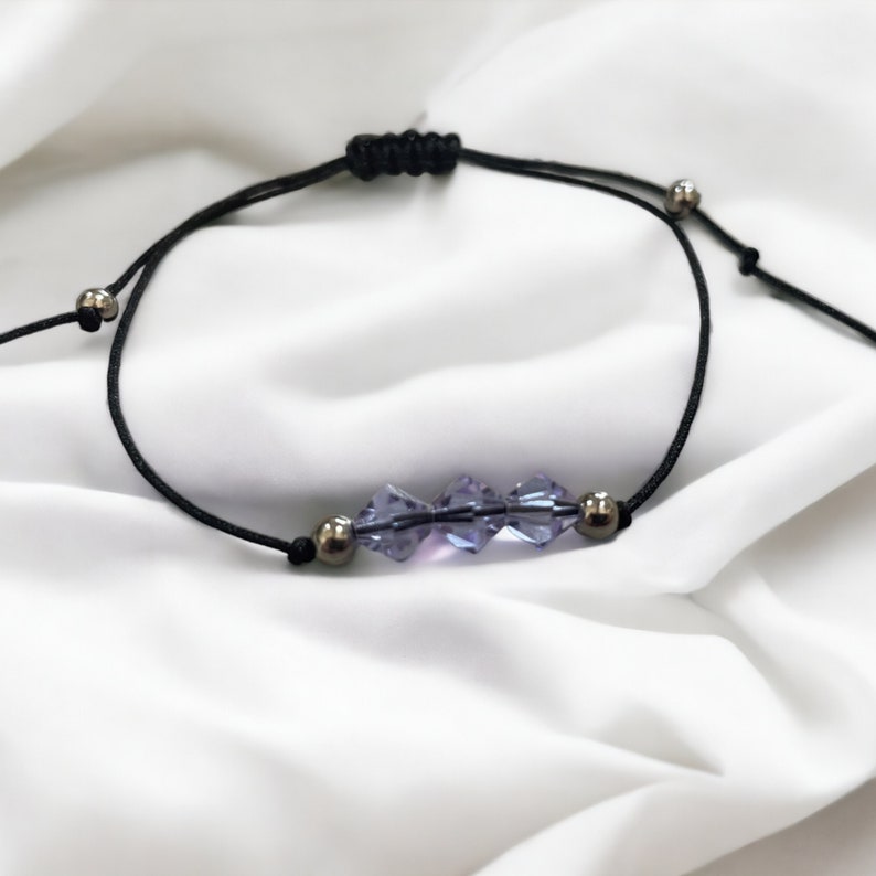 May include: A simple adjustable bracelet with a black cord and silver beads. The bracelet features a row of four purple crystal beads.