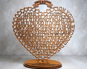 Puede incluir: Decoración de boda en forma de corazón de madera con las palabras "Love is..." talladas. Los nombres "Johnny & Hannah" y la fecha "20.08.25" están en la parte superior. La base tiene un mensaje para la pareja.