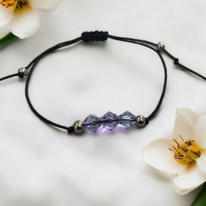 May include: A simple bracelet with a black cord and three purple crystal beads. The bracelet is tied with a knot and has silver metal beads on either side of the crystal beads.