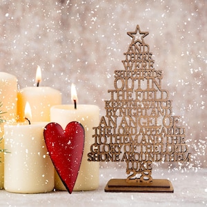 May include: A wooden Christmas tree ornament with the text "And there were shepherds out in the field keeping watch by night and an angel of the Lord appeared to them and the glory of the Lord shone around them. Luke 2:8". The ornament is next to lit candles and a red heart.