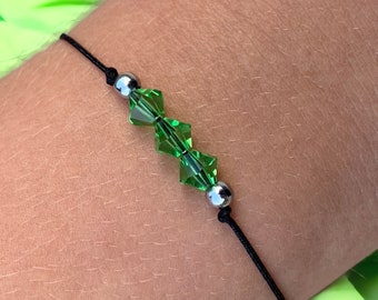 Puede incluir: Una pulsera delicada con tres cristales facetados de color verde esmeralda y dos cuentas plateadas ensartadas en un cordón negro. La pulsera está diseñada para ser ajustable y se muestra en una muñeca.