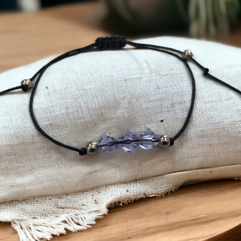 May include: A simple adjustable bracelet with a black cord and a row of small, clear, purple crystal beads.