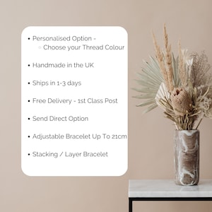 May include: A white card with black text listing the features of a handmade bracelet. The text includes "Personalized Option", "Choose your Thread Colour", "Handmade in the UK", "Ships in 1-3 days", "Free Delivery - 1st Class Post", "Send Direct Option", "Adjustable Bracelet Up To 21cm", and "Stacking / Layer Bracelet". The card is in front of a vase with dried flowers.