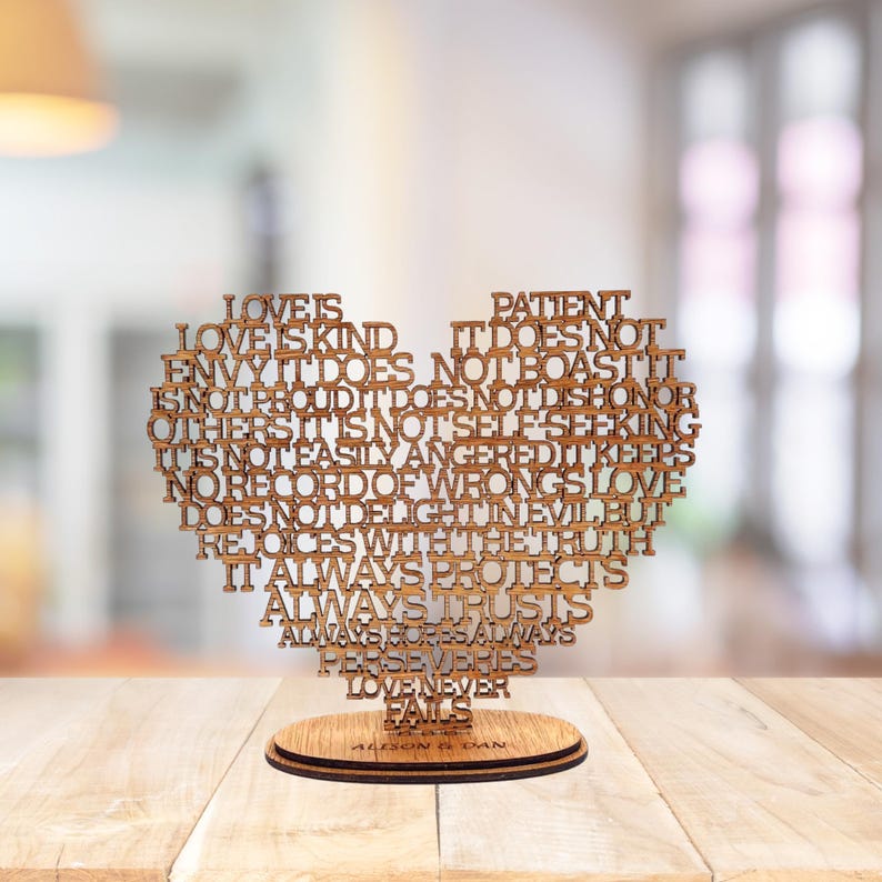 May include: A wooden heart-shaped decorative piece with inspirational text. The heart is made of light brown wood and sits on a round wooden base. The text includes phrases like "Love is Kind" and "Love Never Fails".