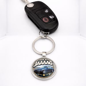 May include: Black car key with a silver key ring attached to a round keychain. The keychain has the text "IT'S A JAAAAAG" above an image of a blue car. The key has buttons for various functions.