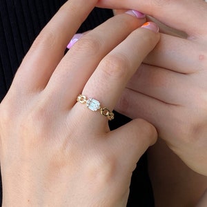 May include: A gold chain ring with a clear emerald-cut gemstone. The ring is on a person's finger.