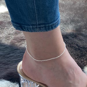 May include: A white pearl anklet on a person's ankle. The anklet is made of small, white pearls that are strung together on a thin chain. The anklet is worn on a person's right ankle, and the person is wearing a pair of clear heeled sandals.