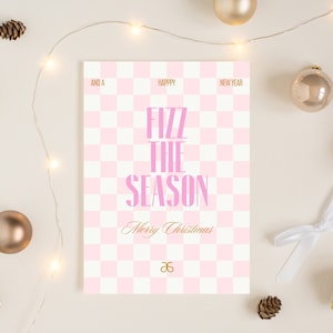 May include: A holiday greeting card with a pink and white checkered pattern. The card reads "Fizz the Season" in pink text, with "Merry Christmas" in gold script. The card is surrounded by gold ornaments, pinecones, and string lights.