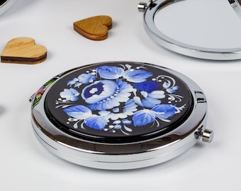 May include: Several round, silver compact mirrors with floral patterns. One mirror is open, showing a reflective surface. The floral designs include red, blue, and white flowers on a black base. Small wooden heart shapes are scattered around the mirrors.