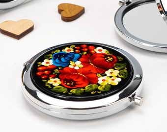 May include: A set of round, silver-toned compact mirrors with black backgrounds and colourful floral designs painted on the lids. The floral designs feature red, blue, yellow, and green flowers.