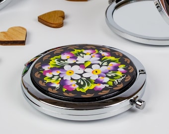 May include: A set of small, round, silver-framed mirrors with painted floral designs. The mirrors are closed and open, showing the reflective surface. The floral designs are in shades of red, white, blue, purple, and green.
