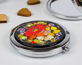 May include: A set of six silver compact mirrors with black backgrounds and colourful floral designs. The mirrors are open and closed, showing the reflective surface and the floral design. The mirrors are surrounded by small wooden hearts.