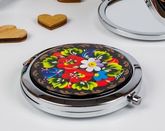 May include: A set of round, silver-toned compact mirrors with black backgrounds and colourful floral designs. The floral designs are painted in red, yellow, blue, green, and white.