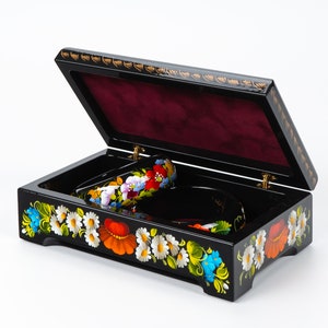 May include: Black wooden box with a hinged lid, decorated with colourful floral patterns. The box is open, revealing a red velvet interior and two decorative items.