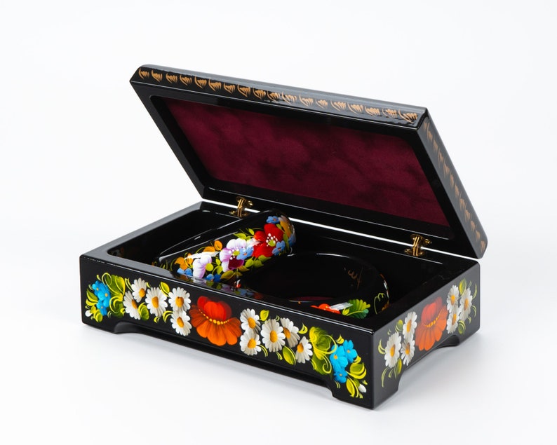 May include: Black wooden box with a hinged lid, decorated with colourful floral patterns. The box is open, revealing a red velvet interior and two decorative items.