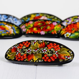 May include: A selection of black, oval hair clips, each decorated with colourful floral patterns. The clips display red berries, yellow and orange flowers, and green leaves. The background is a white wooden surface.